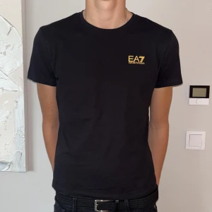 Svart EA7 Emporio Armani t-shirt - Svart t-shirt från EA7 Emporio Armani med gul logga på bröstet och gul detalj på ryggen. Klassisk rund hals och korta ärmar. Snygg och stilren modell i mjuk bomull som passar perfekt till jeans eller shorts.