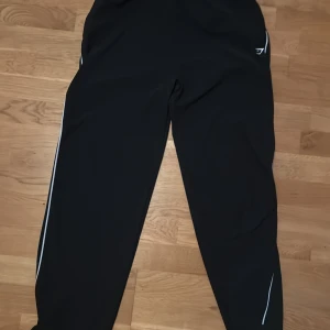Svarta träningsbyxor från Gymshark - Svarta träningsbyxor från Gymshark med vita detaljer längs benen. Byxorna har en sportig look och är tillverkade i ett lätt och stretchigt material som passar perfekt för gymmet eller löprundan.