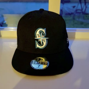 Svart New Era 59FIFTY Seattle Mariners keps - Säljer en svart New Era 59FIFTY keps med Seattle Mariners-logga broderad framtill. Klassisk flat peak och MLB-logga bak. Kepsen är i modellen fitted och har detaljer i vitt, blått och turkost. Perfekt för dig som gillar sportig stil. 