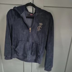 Mörkblå Juicy Couture hoodie i velour - Mörkblå hoodie från Juicy Couture i mjuk velour med huva och dragkedja. Guldiga nitar bildar JC-logga med krona på bröstet och Juicy Couture-text på ryggen. Känguruficka framtill och ribbade muddar. Perfekt för en chill och trendig look.