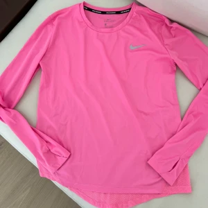 Rosa långärmad träningströja Nike - Nike långärmad träningströja i rosa med meshpanel baktill för extra ventilation. Tröjan har rund halsringning, Nike-logga på bröstet och är tillverkad i lätt, syntetiskt material som andas. Perfekt för löpning eller gymmet.