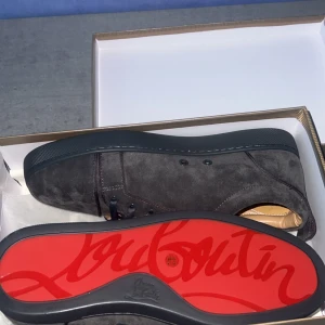 Christian Louboutin svarta mockasneakers - Snygga gråa sneakers från Christian Louboutin i mjuk mocka med klassisk röd sula och diskret logga undertill. Skorna har rund tå, svarta snören och en stilren design som sticker ut med den ikoniska röda undersidan.