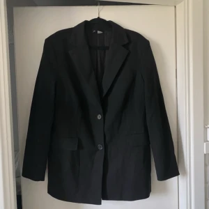 Svart kavaj i polyester och viskos - Stilren svart kavaj med klassisk krage och två knappar framtill. Tillverkad i en mix av polyester, viskos och elastan för en bekväm passform. Kavajen har raka ärmar och två fickor med lock. Perfekt för en snygg och enkel look.