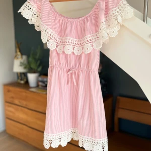 Rosa och vit randig kortklänning xxs - Superfin kortklänning i rosa och vitt med smala ränder, offshoulder och bred volang med vit spetsdetalj i blommönster. Klänningen har knyt i midjan och spetskant längst ner. Perfekt för sommarens alla tillfällen!