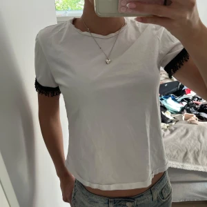Vit topp med svarta spetsdetaljer - Säljer en vit topp med korta ärmar och svarta spetsdetaljer vid ärmsluten. Perfekt för dig som gillar att sticka ut med små detaljer. Passar dig som vill ha en basic topp med en twist.🥰