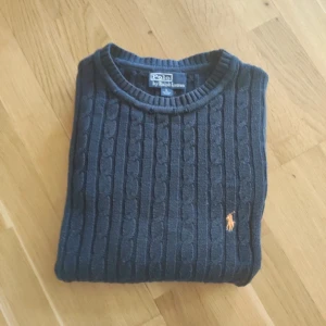 Mörkblå kabelstickad tröja Polo Ralph Lauren - Säljer en mörkblå kabelstickad tröja från Polo Ralph Lauren i storlek L. Tröjan är gjord av 100% pima bomull och har den klassiska broderade Polo-loggan på bröstet i orange. Rund hals och långa ärmar med ribbade muddar för extra stil.