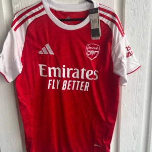 Arsenal matchtröja Gyökeres #14 Adidas - Säljer en officiell Arsenal fotbollströja från Adidas med Gyökeres och nummer 14 på ryggen. Tröjan är röd med vita korta ärmar, har vita detaljer och tryck, samt Champions League-märke på ärmen. Materialet är lätt och andas, perfekt för match eller träning.