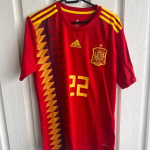 Spanien Isco VM 2018 fotbollströja - Säljer en officiell Spanien fotbollströja från VM 2018 med Isco och nummer 22 på ryggen. Röd med gula detaljer, Adidas-logga och landslagsemblem på bröstet. Snyggt geometriskt mönster på sidan och gula ränder på axlarna. Tillverkad i lätt och ventilerande polyester.