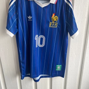 Frankrike Platini #10 Adidas fotbollströja - Retroblå fransk landslagströja från Adidas med vita och röda tunna ränder, klassisk v-ringad krage och korta ärmar. Tryck med nummer 10 och Platini på ryggen, FFF-emblem och Adidas-logga på bröstet. Materialet är lätt och andas, perfekt för match eller samling.