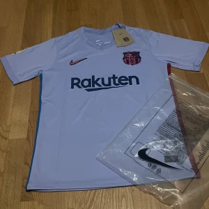 Ljuslila FC Barcelona Nike fotbollströja - Snygg FC Barcelona fotbollströja från Nike i ljuslila med mörkblå Rakuten-tryck och klubbmärke i rosa och mörkblått. Kortärmad modell med V-ringning och Dri-FIT-material som andas. Perfekt för dig som vill ha en unik Barca-tröja med modern look.