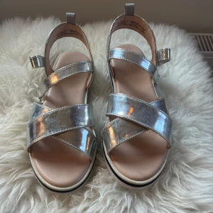 Silvriga sandaler från H&M - Snygga sandaler från H&M i metallic silver med breda korslagda remmar och justerbar kardborre vid ankeln. Platt vit sula med svart detalj och öppen tå. Perfekt för dig som vill sticka ut med glansiga detaljer i sommar. Storlek 36 och använda endast en gång💓