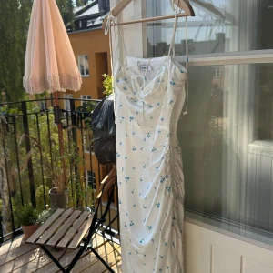 Vit blommig långklänning från Adoore - Supersöt långklänning från Adoore i vit med små blå blommor. Klänningen har tunna axelband med knyt, draperad front och lätt rynkade sidor som ger en snygg siluett. Perfekt för varma dagar och somriga vibes. OBS klänningens har en trasig söm på ena sidan, det syns inte jättemke och skulle vara enkelt att laga om man vill det! 