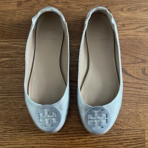 Silver ballerinaskor från Tory Burch - Snygga ballerinaskor från Tory Burch i metallic silver med rund tå och platt sula. Skorna har en stor logotyp i silver framtill och är tillverkade i skinn med mjuk insida. Perfekta för dig som gillar stilrena detaljer och vill ha något som sticker ut. DE ÄR ÄKTA!