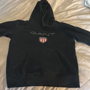 Svart hoodie från GANT med logga - Svart hoodie från GANT med stor broderad logga och sköld på bröstet. Klassisk känguruficka framtill och huva. Snygga detaljer med röd, vit och blå rand på insidan av kragen. priset kan diskuteras