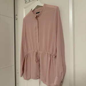 Ljusrosa skjorta från Gina Tricot - Säljer en ljusrosa skjorta från Gina Tricot med lång ärm och knappar framtill. Skjortan har en rund halsringning och snörning i midjan för en snygg passform. Materialet är mjukt och lätt, perfekt för en chill look. Storlek 42 men liten i storleken.