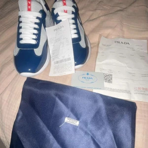 Blå och vita sneakers från Prada - Säljer ett par snygga Prada sneakers i blått och vitt med röda detaljer på plösen och sulan. Skorna har en rund tå, vita snören och meshpaneler för extra ventilation. Snygg kontrast mellan blankt blått material och vit mesh. Kommer med originalpåse och äkthetsbevis.