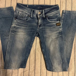 G-Star ljusgråblå jeans – strl 24/32 - Snygga blå jeans från G-Star RAW strl 24/32 med coola slitningar och detaljerade sömmar. Baktill finns dragkedjor på fickorna och en svart patch med logga. Jeansen har normal passform och raka ben, tillverkade i klassiskt jeanstyg. Perfekt för dig som gillar streetstyle.