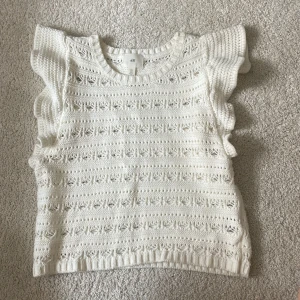 Vit stickad topp med volang från H&M - Supersöt vit stickad topp från H&M med volangdetaljer på axlarna och ett snyggt hålmönster över hela plagget. Toppen är ärmlös och har en rund halsringning. Perfekt för dig som gillar en romantisk och chill stil.