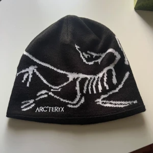 Svart mössa Arc'teryx med vit grafik - Svart mössa från Arc'teryx med coolt vitt dinosaurieskelett-motiv och broderad logga framtill. Modellen heter Fernie Toque och är tillverkad i 100% akryl. Snygg, enkel passform som sitter skönt på huvudet och passar perfekt när det är kallt ute.