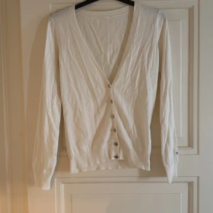 Vit kofta från GANT - Vit kofta från GANT med v-ringning och silvriga knappar framtill. Långärmad och ribbad vid muddar och nederkant. Broderad GANT-logga på ärmen. Mjuk och tunn i bomull, perfekt för lager-på-lager. Enkel och clean stil som funkar till vad som! 