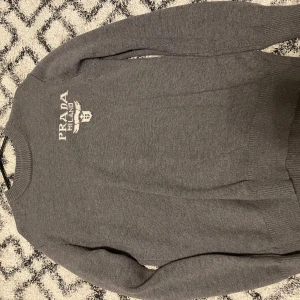Grå stickad tröja från Prada - Säljer en snygg grå stickad tröja från Prada med broderad logga på bröstet. Tröjan har rund hals och långa ärmar, perfekt för dig som gillar stilrena designer och exklusiva märken. Passar dig som vill ha en klassisk look med lyxig känsla.