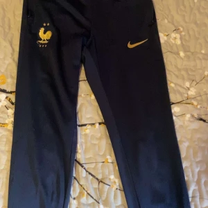 Nike blå träningsbyxor Frankrike Slim - Säljer ett par mörkblå träningsbyxor från Nike med Frankrikes landslagsmärke och guldfärgad swoosh. Slim fit-modell med Dri-Fit-material som andas och ger skön passform. Perfekta för fotboll eller annan träning. Snyggt guldtryck på låret.