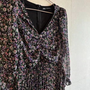 Blommig långklänning från Zara - Supersöt långklänning från Zara med svart botten och blommigt mönster i lila, rosa och grönt. Klänningen har v-ringning, långärmade transparenta ärmar och plisserad kjol.