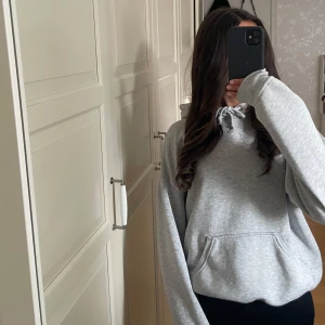 Grå hoodie med snörning och ficka - Säljer en enkel grå hoodie från Bikbok med huva och snörning framtill. Tröjan har en stor magficka och ribbade muddar vid ärmslut och nederkant. Perfekt för chill dagar och har en loose passform. Materialet känns mjukt och är troligen bomull eller polyester.