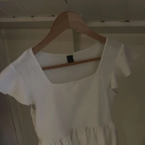 Vit kortklänning med volangärmar - Säljer en söt vit kortklänning med volangärmar och smock upptill. Klänningen har en fyrkantig ringning och är tillverkad i ett lätt, strukturerat tyg som ger en fräsch och somrig känsla. Perfekt för dig som gillar enkel och stilren design.