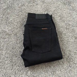  Nudie Jeans Co, 26/34 - Säljer ett par svarta jeans från Nudie Jeans Co i storlek 26/34. Modellen har klassisk femficksdesign, svarta sömmar och silverfärgade nitar. Jeansen är raka i snittet och har en snygg, stilren look med svart läderpatch bak i midjan.
