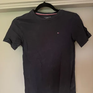 Svart t-shirt Tommy Hilfiger - Svart t-shirt från Tommy Hilfiger med klassisk rund hals och liten broderad logga på bröstet. T-shirten har korta ärmar och är gjord i mjuk bomull. Perfekt för dig som gillar stilrena och enkla plagg med en touch av märkesstil.