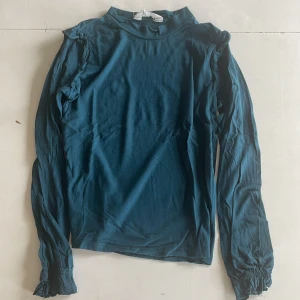 Mörkblå långärmad topp med volang - Snygg mörkblå långärmad topp från H&M med volangdetaljer längs axlarna och lätt rynkade ärmslut. Materialet är mjukt och stretchigt, perfekt för dig som gillar en enkel men ändå unik look. Passar dig som vill ha något stilrent med en twist.