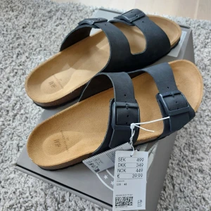 Premium Quality - Svarta lädersandaler från H&M - Säljer ett par svarta sandaler från H&M Premium Selection i äkta läder. Sandalerna är helt oanvända. Modellen har två breda remmar med stora svarta spännen och en mjuk, beige innersula. Perfekt för dig som gillar enkel och stilren design med sköna material.