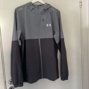 Grå vindjacka från Under Armour - Snygg tvåtonad grå vindjacka från Under Armour med huva och hel dragkedja framtill. Jackan har lång ärm, elastiska muddar med logga och en bröstficka med dragkedja. Perfekt för träning eller när du vill ha en sportig look. Materialet är lätt och syntetiskt. KÖPT FÖR 750