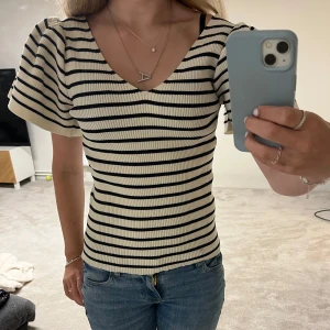 Randig ribbad topp från Vero Moda - Säljer en snygg ribbad topp från Vero Moda i storlek M. Toppen är vit med mörkblå/svart ränder, har v-ringad hals och korta puffärmar. Super fint skick! 