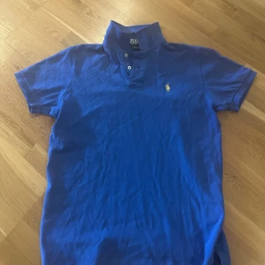 Blå Polo Ralph Lauren t-shirt - Snygg blå t-shirt från Polo Ralph Lauren med klassisk pikékrage och tre knappar framtill. Ikoniska broderade loggan i gult på bröstet. Modellen är custom fit och har korta ärmar. Perfekt för dig som gillar stilren och sportig look.