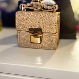 Beige AirPod väska  från iDeal of Sweden - Snygg liten handväska i beige med krokodilpräglat mönster och guldfärgade detaljer. Väskan har ett stilrent fyrkantigt format, ett flätat handtag och ett elegant lås framtill. Perfekt för att bära småsaker och airpods i.