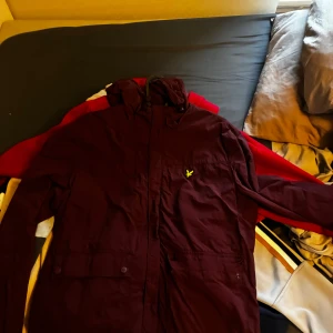Vinröd vindjacka från Lyle & Scott - Snygg vinröd vindjacka från Lyle & Scott med huva och gul logga på bröstet. Jackan har långa ärmar, två fickor med knappar och dragkedja framtill. Perfekt för dig som vill ha en stilren jacka med sportig vibe.