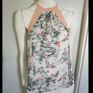 Mönstrad ärmlös blus med persikofärgad hals - Snygg blus med persikofärgad halsringning och asiatiskt inspirerat mönster med fåglar, blommor och vatten. Blusen är ärmlös och har ett lätt, transparent yttertyg med vitt foder under. 