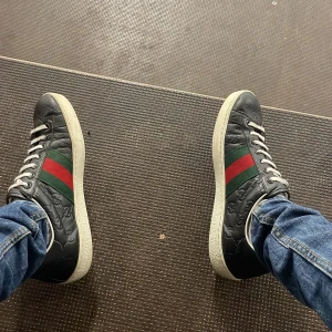 Gucci svarta sneakers med röd/grön rand - Säljer ett par svarta Gucci sneakers i skinn med klassiska röd och grön rand på sidan. Skorna har vit platt sula, vita snören och präglat GG-mönster i lädret. Snygg och tidlös modell som levereras med originalkartong och dustbag.