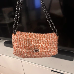 Rutig axelväska med kedja från Becksöndergaarden  - Snygg rutig axelväska från Becksöndergaarden i orange och beige med grov textilstruktur. Väskan har en cool kedjerem i metall och ett litet metallspänne framtill. Insidan är fodrad med svart tyg och har en praktisk innerficka. Perfekt för att lyfta din outfit.