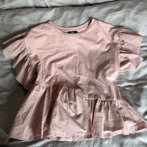 Rosa volangblus från C&A - Supersöt ljusrosa blus från C&A i mjuk bomull. Blusen har volanger både på ärmarna och nedtill, vilket ger en extra feminin och lekfull vibe. Rund halsringning och korta ärmar med volangdetaljer gör den perfekt för vår och sommar.