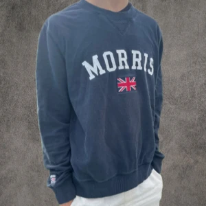 Svart sweatshirt från Morris - Snygg svart sweatshirt från Morris med broderad logga och brittisk flagga på bröstet. Tröjan har rund hals, ribbade muddar och en patch på ärmen. Perfekt för dig som gillar klassisk och sportig stil.