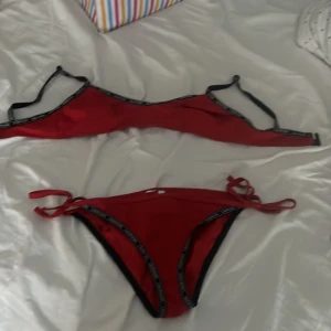 Röd bikini från Calvin Klein med svarta detaljer - Snygg röd bikini från Calvin Klein med svarta kanter och logotypband. Toppen har smala axelband och bikinitrosan har knyt i sidorna för perfekt passform. Materialet är mjukt och stretchigt, perfekt för stranden eller poolen. Stilren och sportig look!