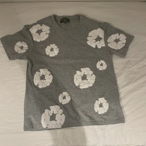 Grå t-shirt med vita tryck från Denim Tears - Säljer en grå t-shirt från Denim Tears i storlek S. T-shirten har coola vita tryck i form av cirklar över hela plagget och klassisk rund hals. Materialet är mjuk bomull och passformen är normal. Perfekt för dig som vill sticka ut med en unik design.