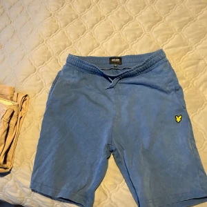 Blå shorts från Lyle & Scott - Blå mjuka shorts från Lyle & Scott med gul logga på vänster ben. De har snörning i midjan och är tillverkade i bomull, vilket gör dem sköna och avslappnade. Perfekta för chill dagar eller träning. Klassisk passform och enkel stil.