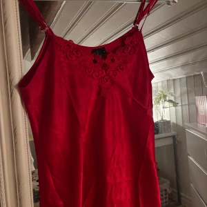 Röd kort lattlinne med spetsdetaljer - Säljer ett rött nattlinne med smala axelband och snygga spetsdetaljer vid halsringningen och nederkanten. Klänningen är kort och har en rak passform, Materialet känns mjukt och har en lätt glans.