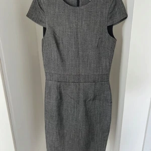 Grå kortärmad klänning  - Snygg grå klänning med diskret rutig struktur och korta ärmar. Modellen är figurnära med rund hals och markerad midja, perfekt för en stilren look. Materialet känns mjukt och bekvämt, och klänningen har en klassisk siluett som passar till många tillfällen.