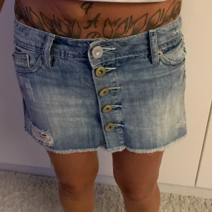 Blå jeanskjol med knappar och slitningar - Säljer en kort jeanskjol från Gini i ljusblå tvätt med slitna detaljer och fransad kant. Kjolen har fem knappar framtill, fickor både fram och bak samt bälteshällor. Perfekt för dig som gillar en avslappnad och cool stil..  midjamått: 38cm