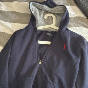Mörkblå hoodie från Polo Ralph Lauren - Snygg mörkblå hoodie från Polo Ralph Lauren med röd broderad logga på bröstet. Tröjan har huva, dragkedja framtill och två fickor. Insidan är mjuk och skön, perfekt för chill dagar. Passar dig som gillar klassisk streetstyle.